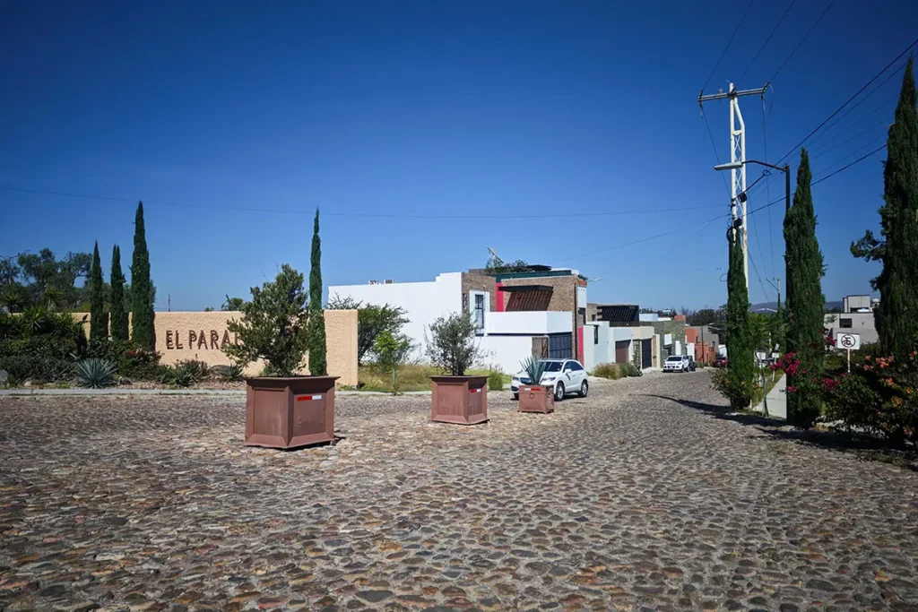 Cobblestone street at the entrance of El Paraíso in San Miguel de Allende, featuring the neighborhood sign, tall cypress trees, and modern residential homes under a clear blue sky.