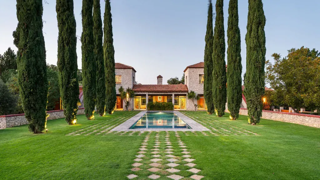 Luxury hacienda for sale in Mexico featuring traditional stone architecture, terracotta roof, twin towers, reflection pool, manicured gardens with towering cypress trees at dusk