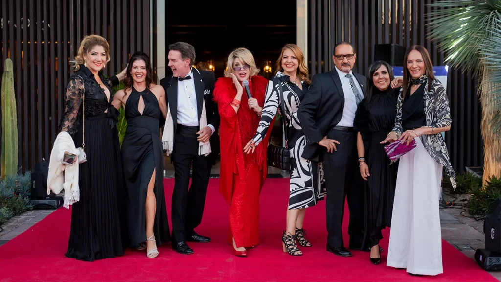 Group of eight adults in formal attire posing on a red carpet outside a modern venue.