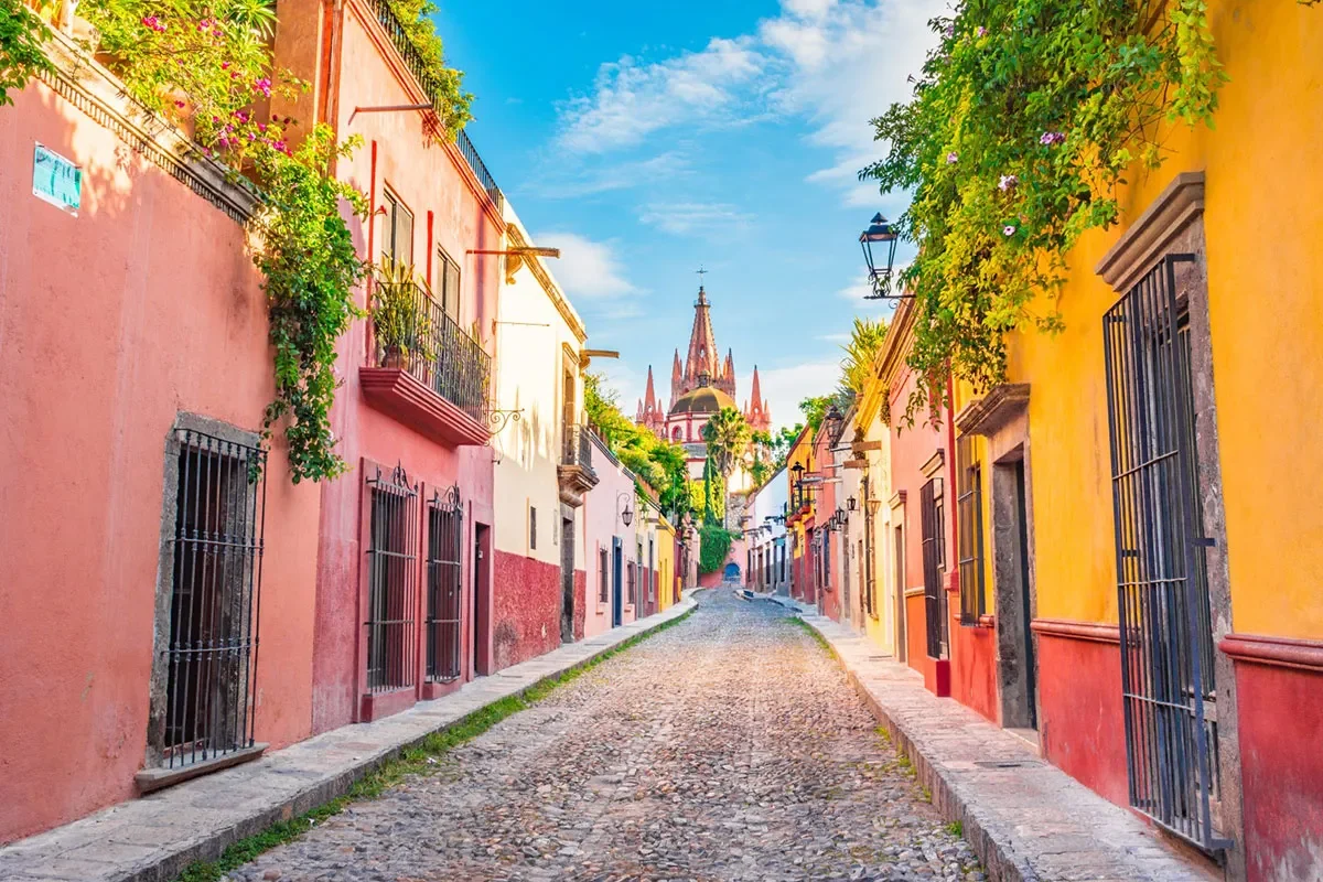 Colorful cobblestone street in San Miguel de Allende with view of Parroquia de San Miguel Arcángel in the distance, reflecting the vibrant local lifestyle and historic charm.